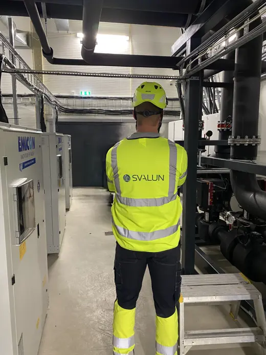 Driftsingeniør som står i en energisentral med gule arbeidsklær med Svalun logo på ryggen.